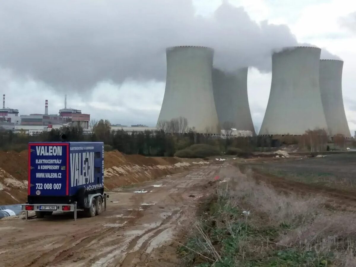Modrá mobilní elektrocentrála Valeon na přívěsu u staveništní cesty, v pozadí chladicí věže jaderné elektrárny
